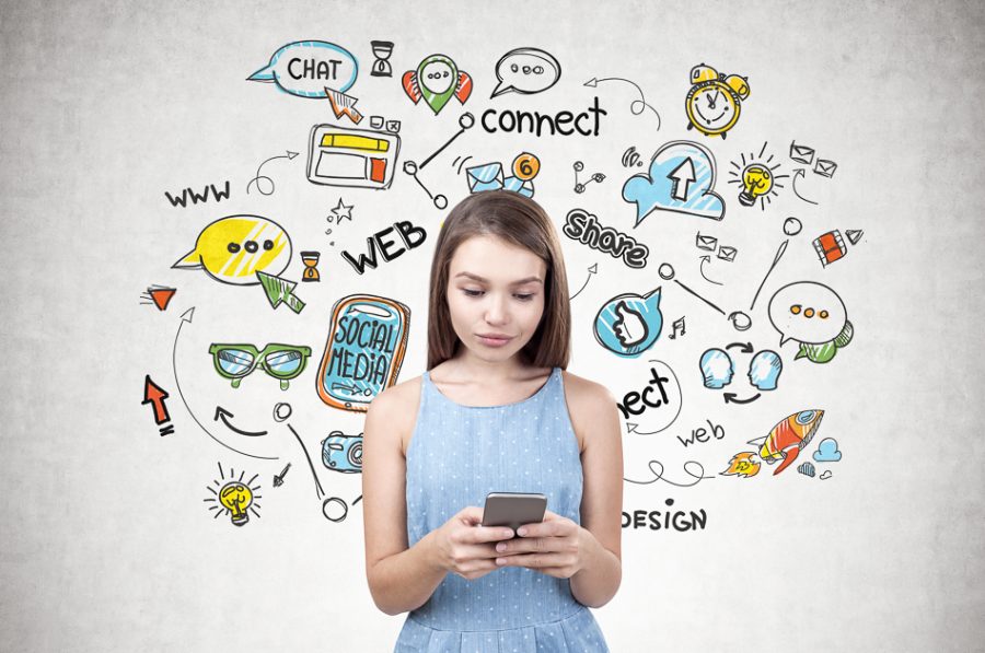 Young woman with long fair hair wearing blue dress holding smartphone with both hands and looking at the screen. Concrete wall with colorful social media icons